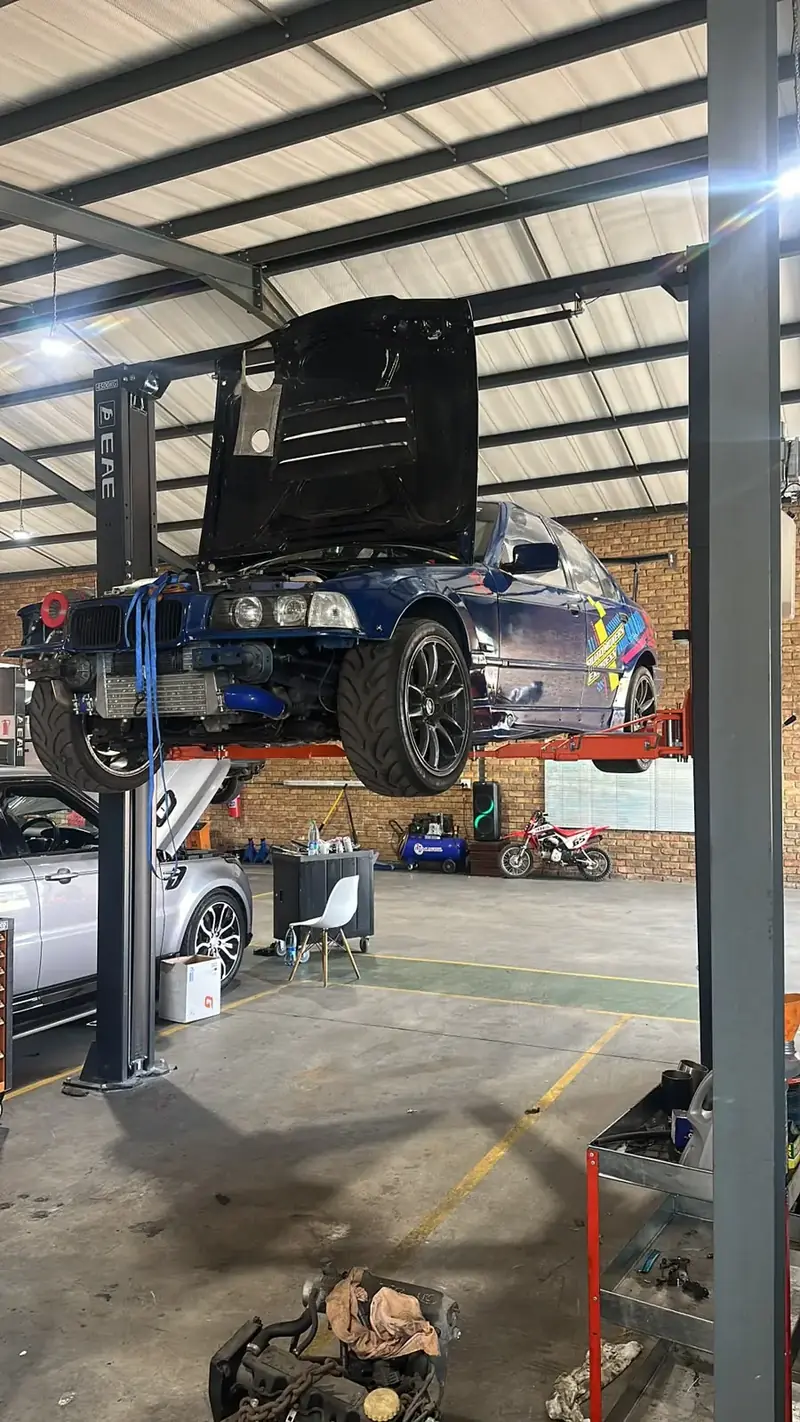 Blue BMW E36 track car on a two-post lift with the bonnet open, showing a front-mount intercooler and blue silicone hoses
