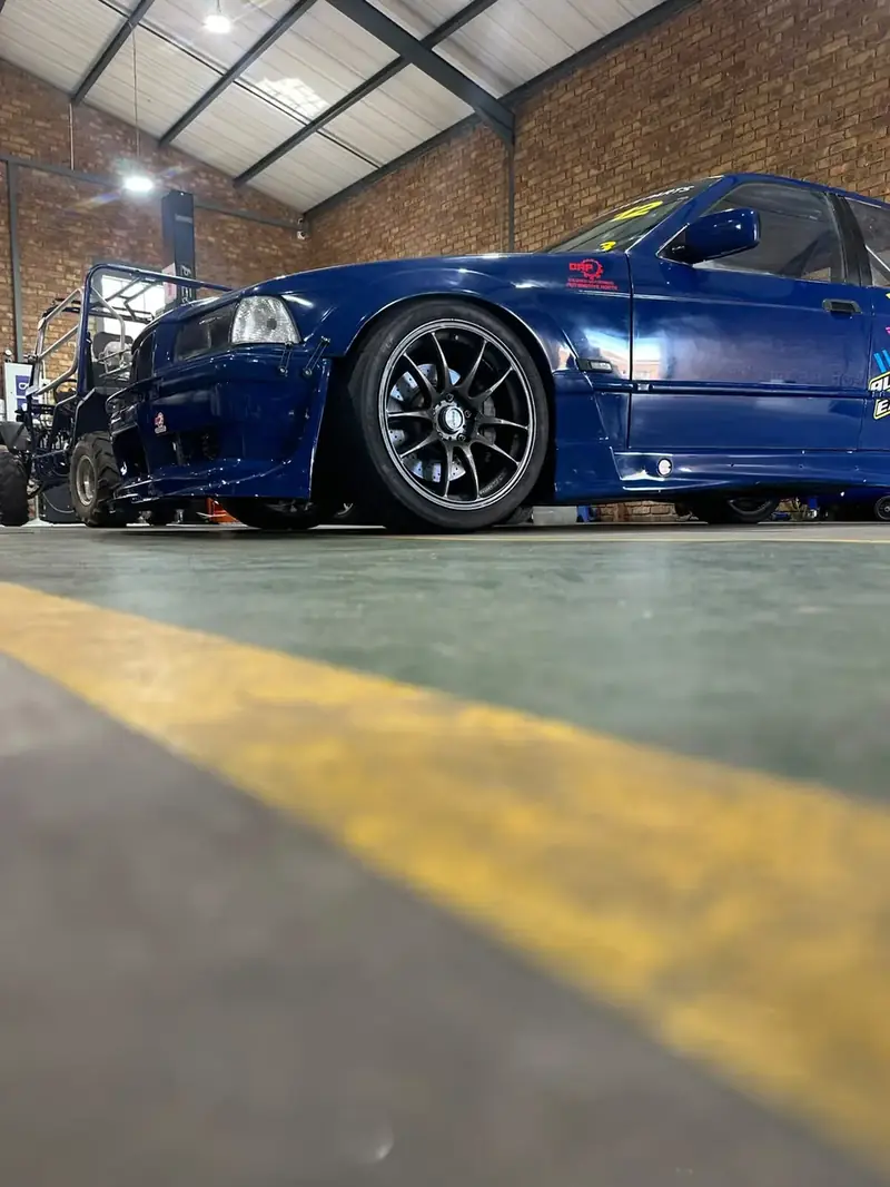 Low-angle view of the BMW E36 in the workshop, showing its wide semi-slick tyres and cross-drilled brake disc