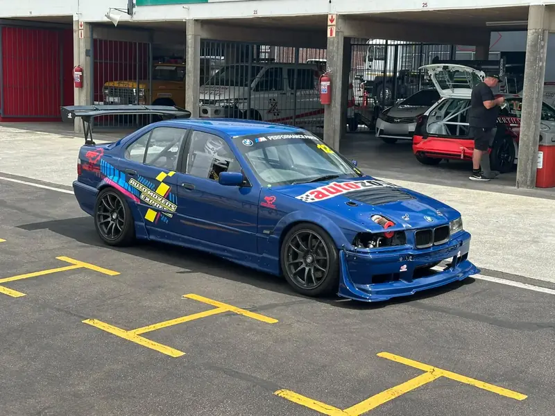 Blue BMW E36 race car number 12 in a pit lane, with a large rear wing, front splitter, roll cage, DAP logo on the rear door and the driver in a helmet